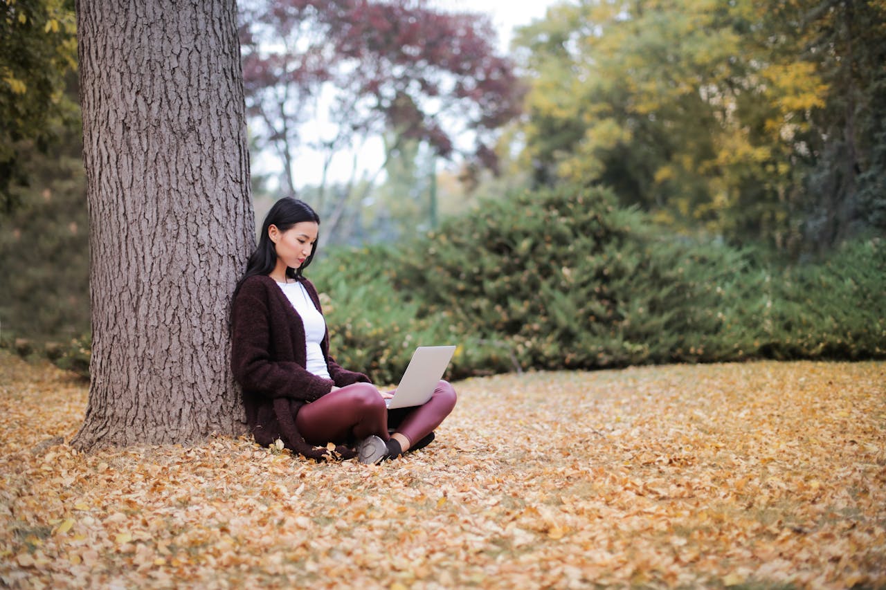 A young woman using a laptop under a tree in a park during fall.