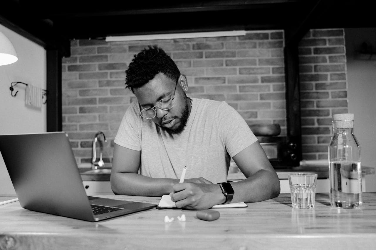 African American man working from home, writing at a desk with a laptop. Monochrome setting enhances focus.