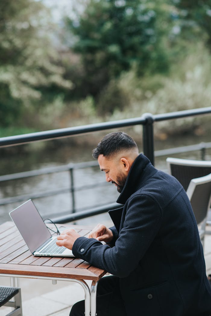 Man working on a laptop at an outdoor café overlooking a scenic river.